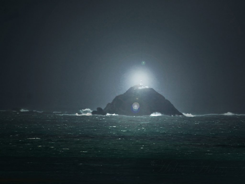 A lighthouse lit up at night time on an isolated rock in a rough sea.