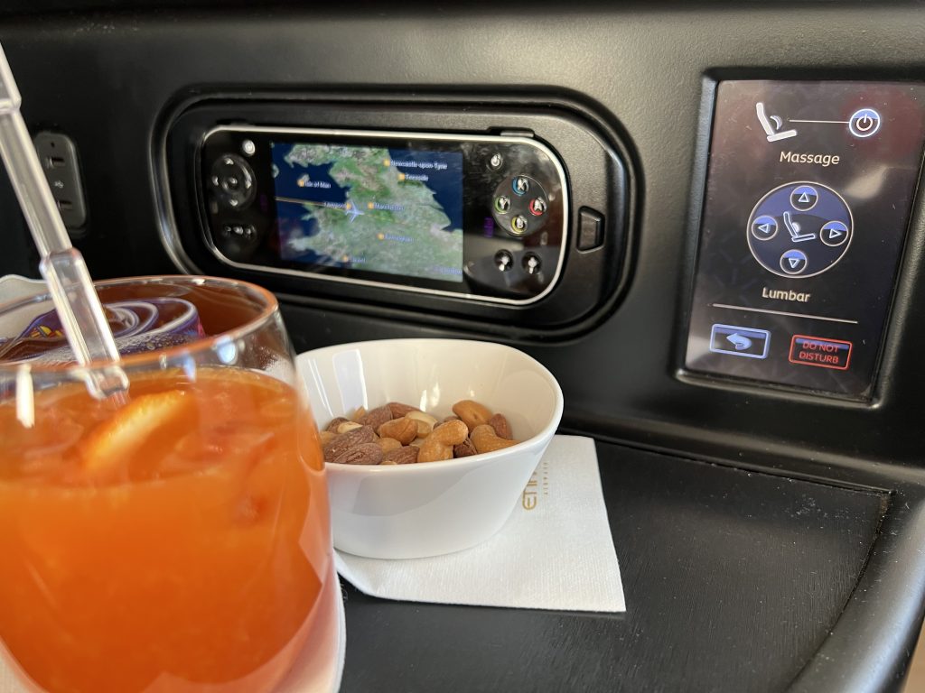 From an aircraft business class seat, a glass of Campari, nut, a tv controller and a sign indicating seat settings including massage
