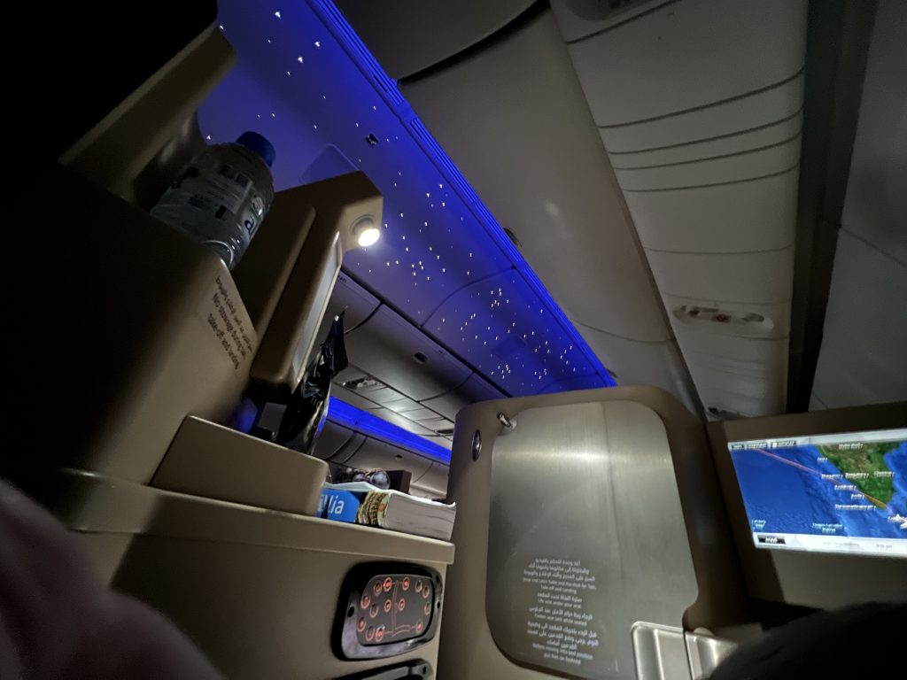 From an aircraft business class seat, lying down, the reading light and beyond the ceiling light is a scattering of pin-prick lights made to look like stars against a blue background