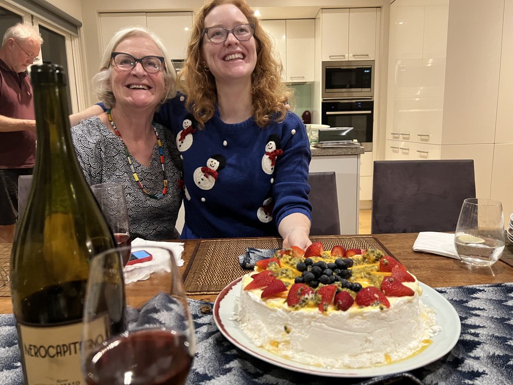 In the foreground table clutter but centrally a beautiful Pavlova decorated with strawberries, passion fruit and blueberries, offered by cook Elise with her other  arm around Jenny her mother with father John in the background.