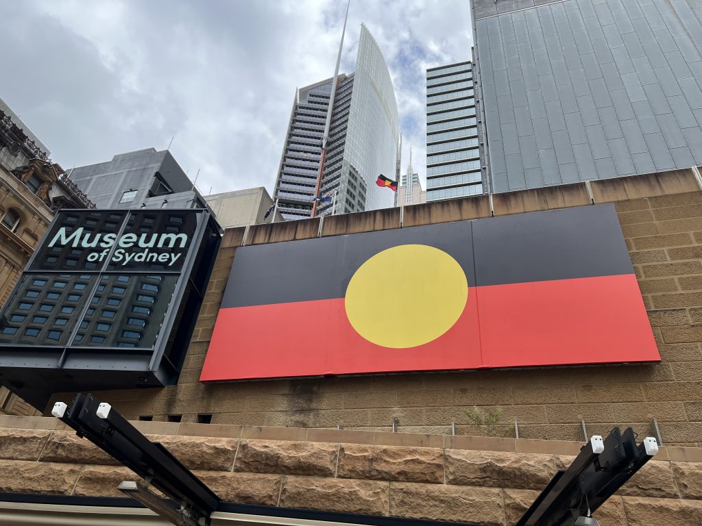In the foreground the Aboriginal flag and sign for the Museum of Sydney, behind the hi-rise buildings of the Sydney Central Business District.