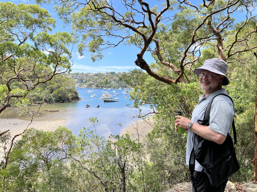 John standing and smiling at the camera as we are overlooking a tree line inlet of the sea with a sandy shore and boats at mooring in the middle distance, and in the far distance, expensive houses across a larger inlet.