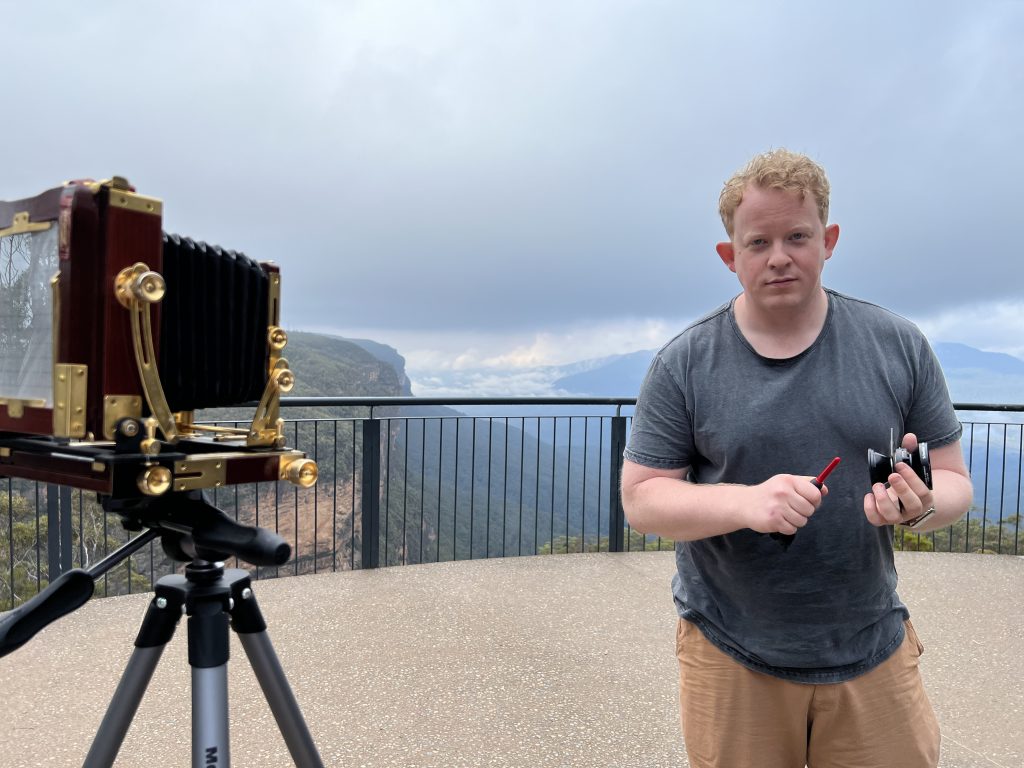 An old-fashioned camera and Patrick stand on a lookout platform, behind the rail is a view over a valley in the Blue Mountains.