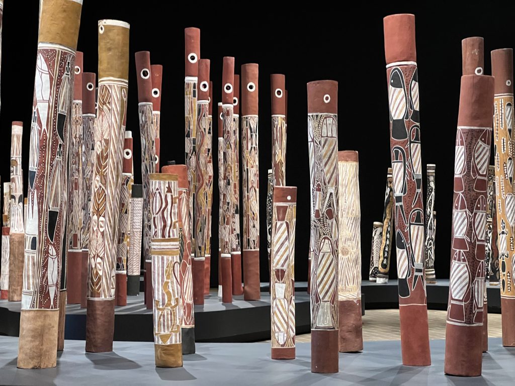 An array of tall decorated vertical pipes painted with ochre, black and white, exhibited with a black background.