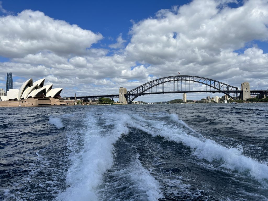 The wake of a speeding boat. IN the distance the Sydney Harbour Bridge and the Opera House under a sunny blue sky with white clouds.
