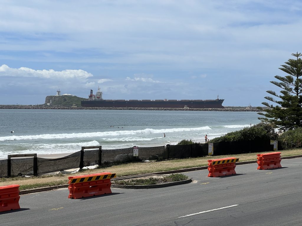 A large boat is entering a harbour in the distance, in the middle distance are waves breaking on a beach and nearer are barriers to keep cars safe from an eroding road.