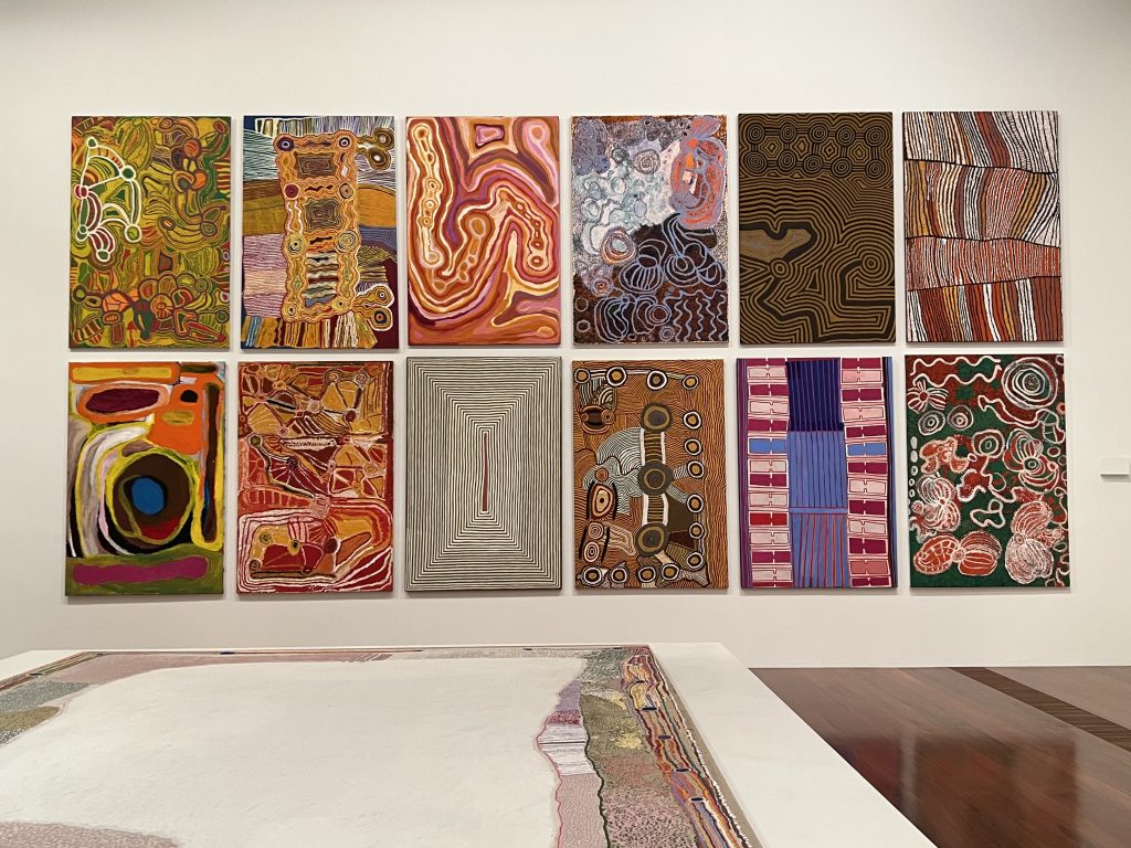 In the foreground is a large horizontal canvas, and hanging on the wall two rows of six indigenous paintings are juxtaposed.