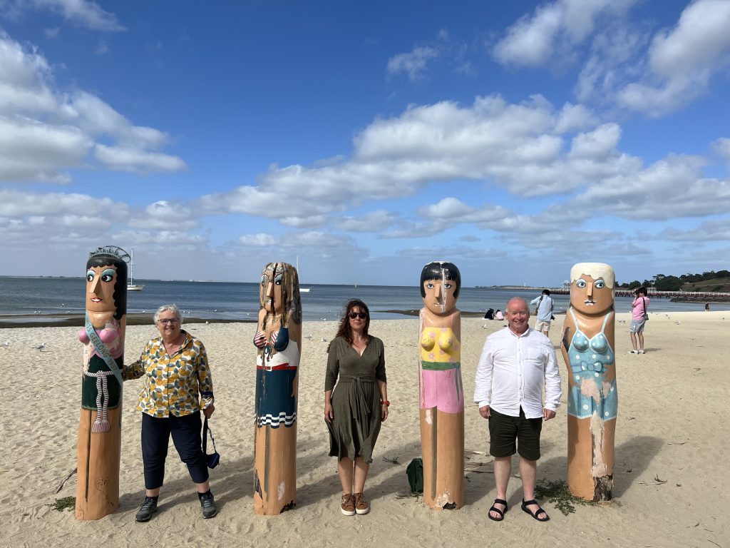 Three people standing on a beach under blue sunny skies between three wooden totems decorated to look like four women.