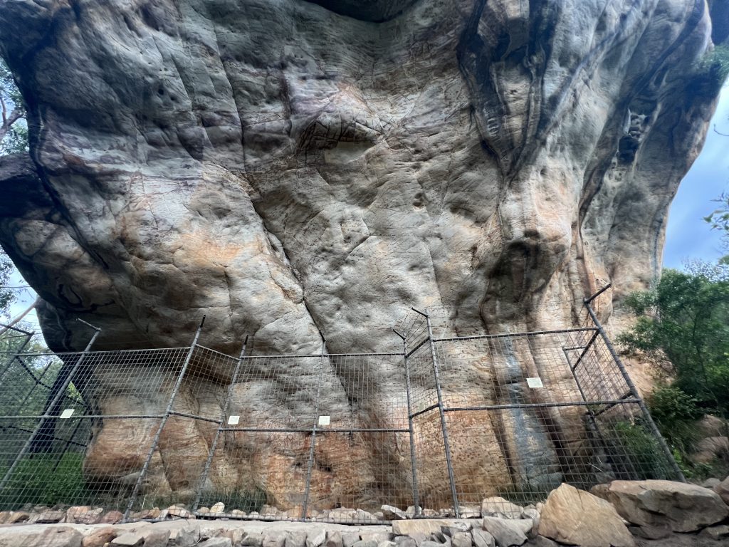 A massive overhanging rock, at the foot a fenced off area behind which is Aboriginal rock art.