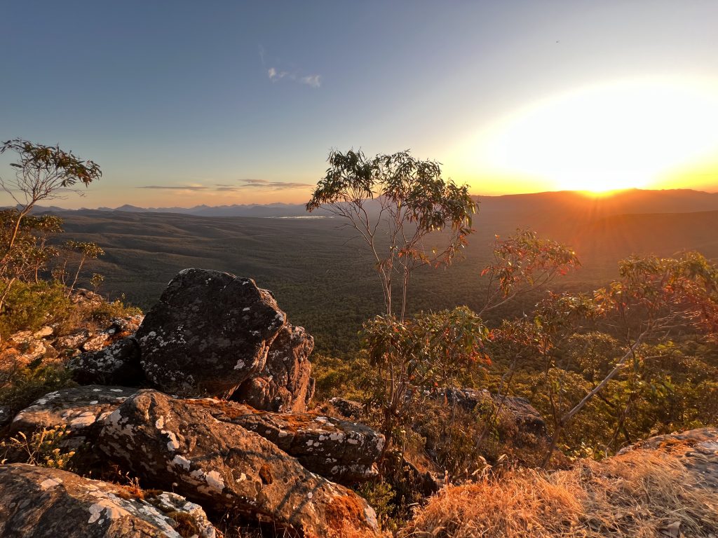 A photograph of Gariwerd / Grampian National Park in Victoria, Australia with boulders and bush in the foreground and distant sawtooth mountains and a brilliant setting sun