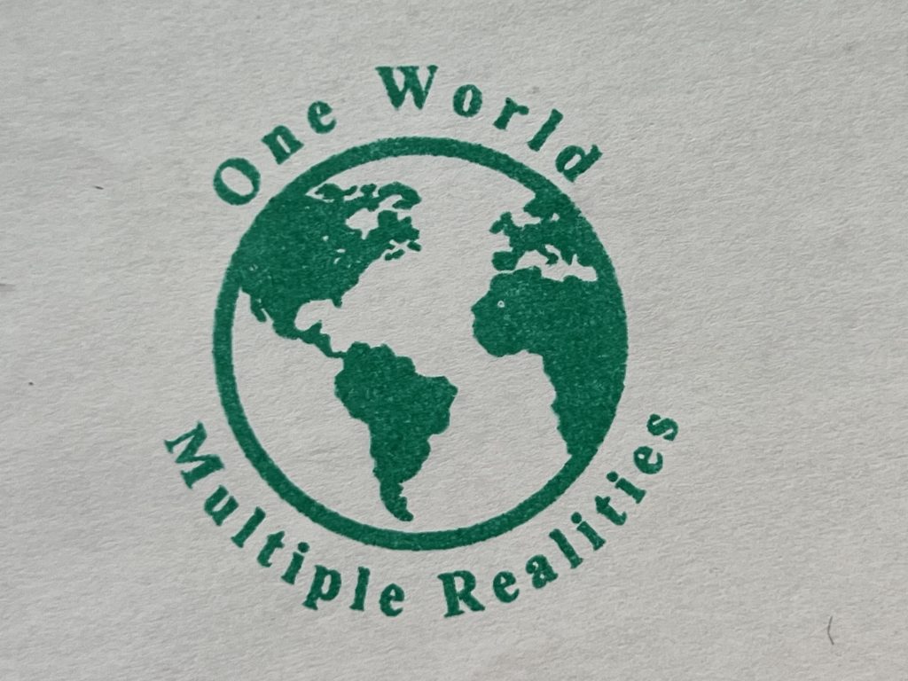 A stamp of an image of the world with the text 'One World - Multiple Realities' arranged around as a circle