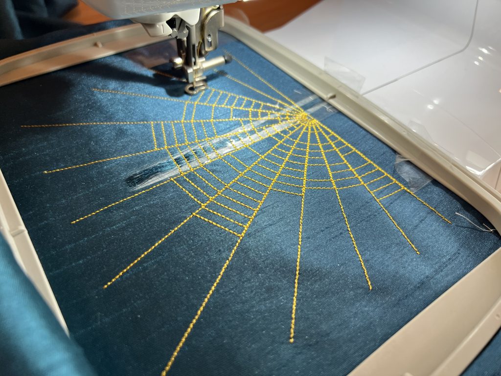 Material in an embroidery machine frame being stitched with lines of thread radiating from a point and cross-connecting lines to look like a spider's web.