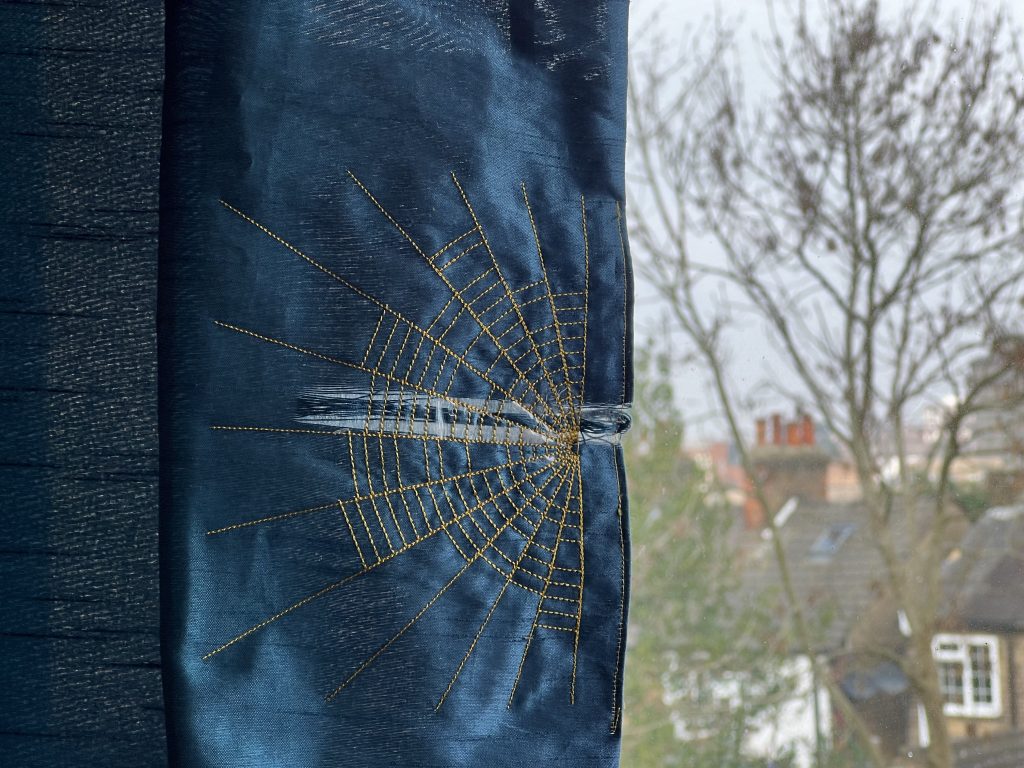 Material finished being stitched with lines of thread radiating from a point and cross-connecting lines to look like a spider's web hanging in a window.