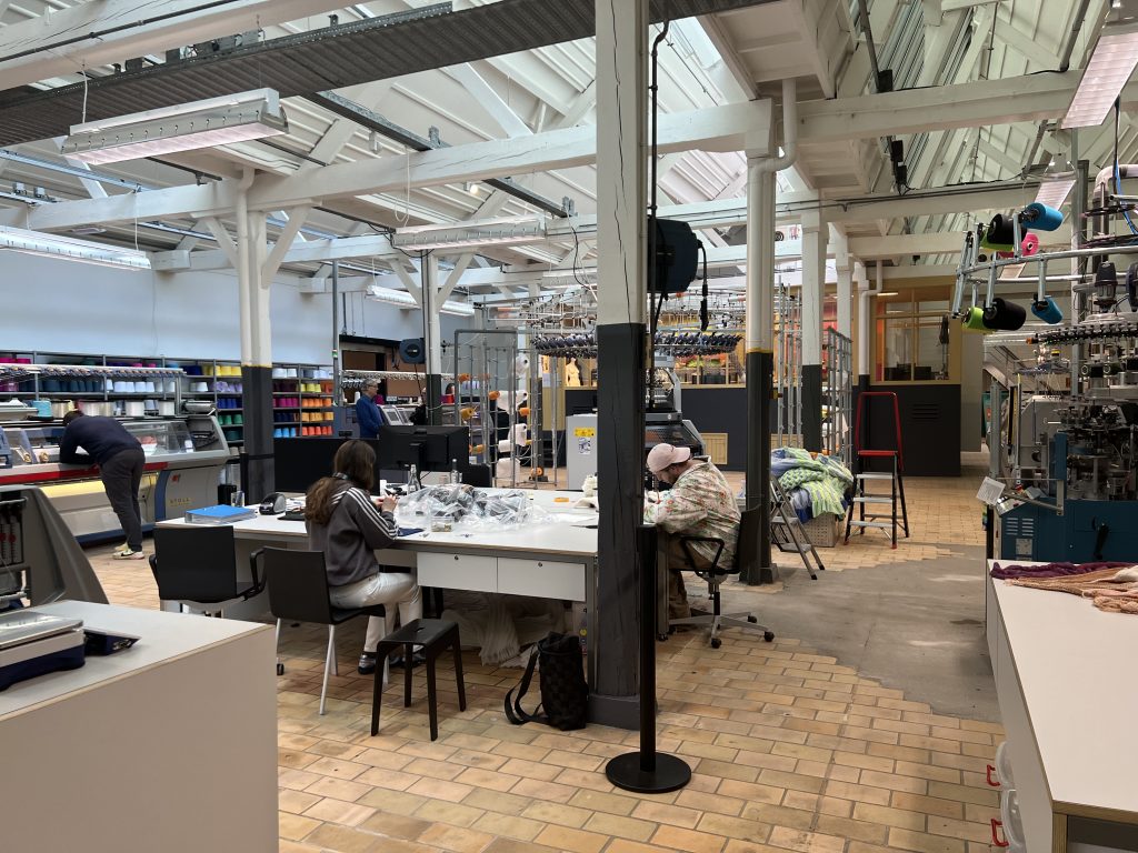 A textile workshop, with artists working at a table in the foreground and machinery and colourful bobbins of thread in the background.